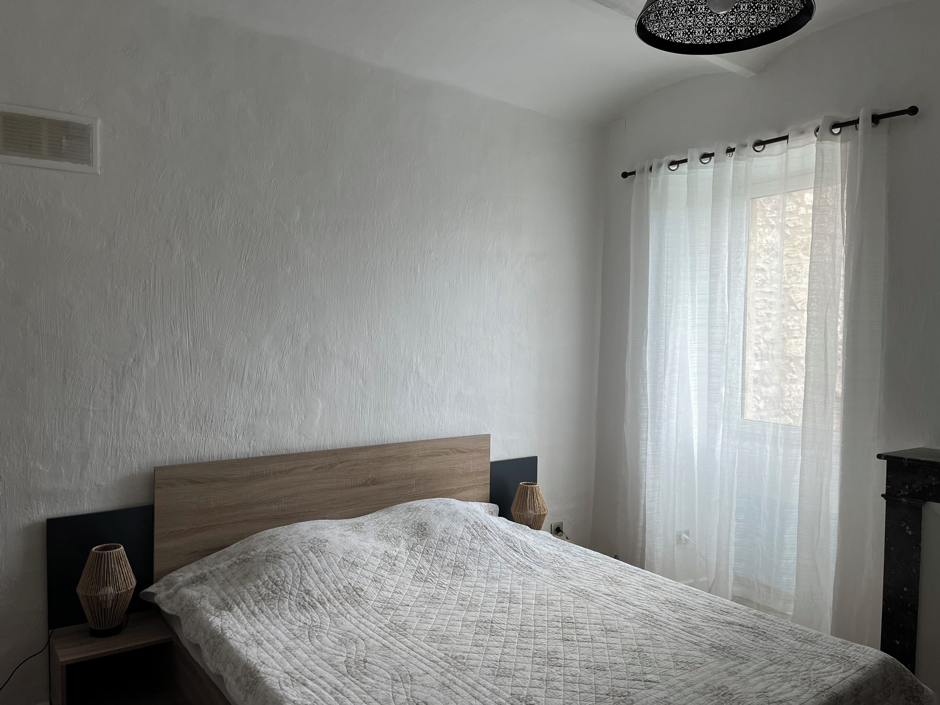 Una camera da letto con una testiera in legno, una coperta trapuntata, un comodino con una lampada e tende leggere su una finestra.