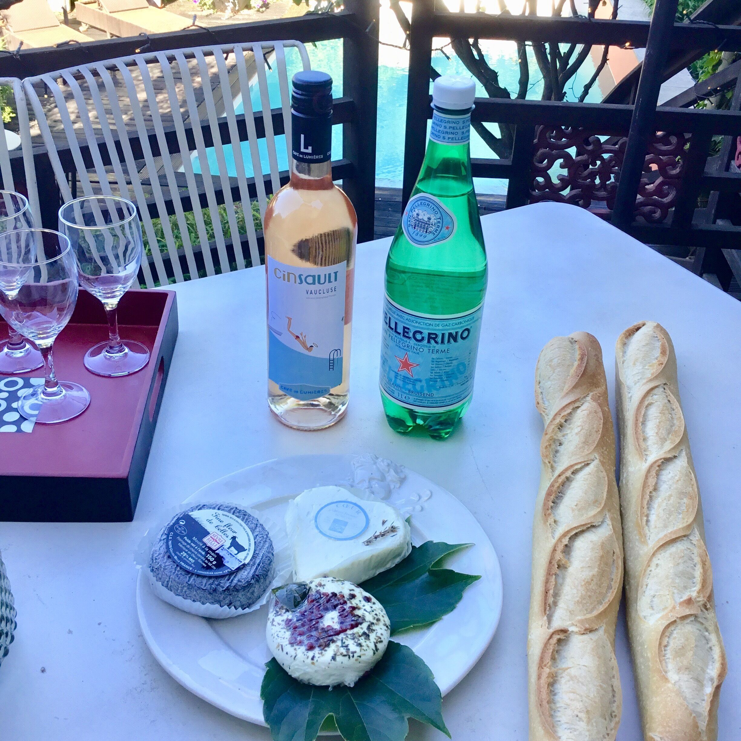A table with a bottle of Cinsault wine, a bottle of Pellegrino water, two glasses, a plate with cheese and bread, and a white chair.