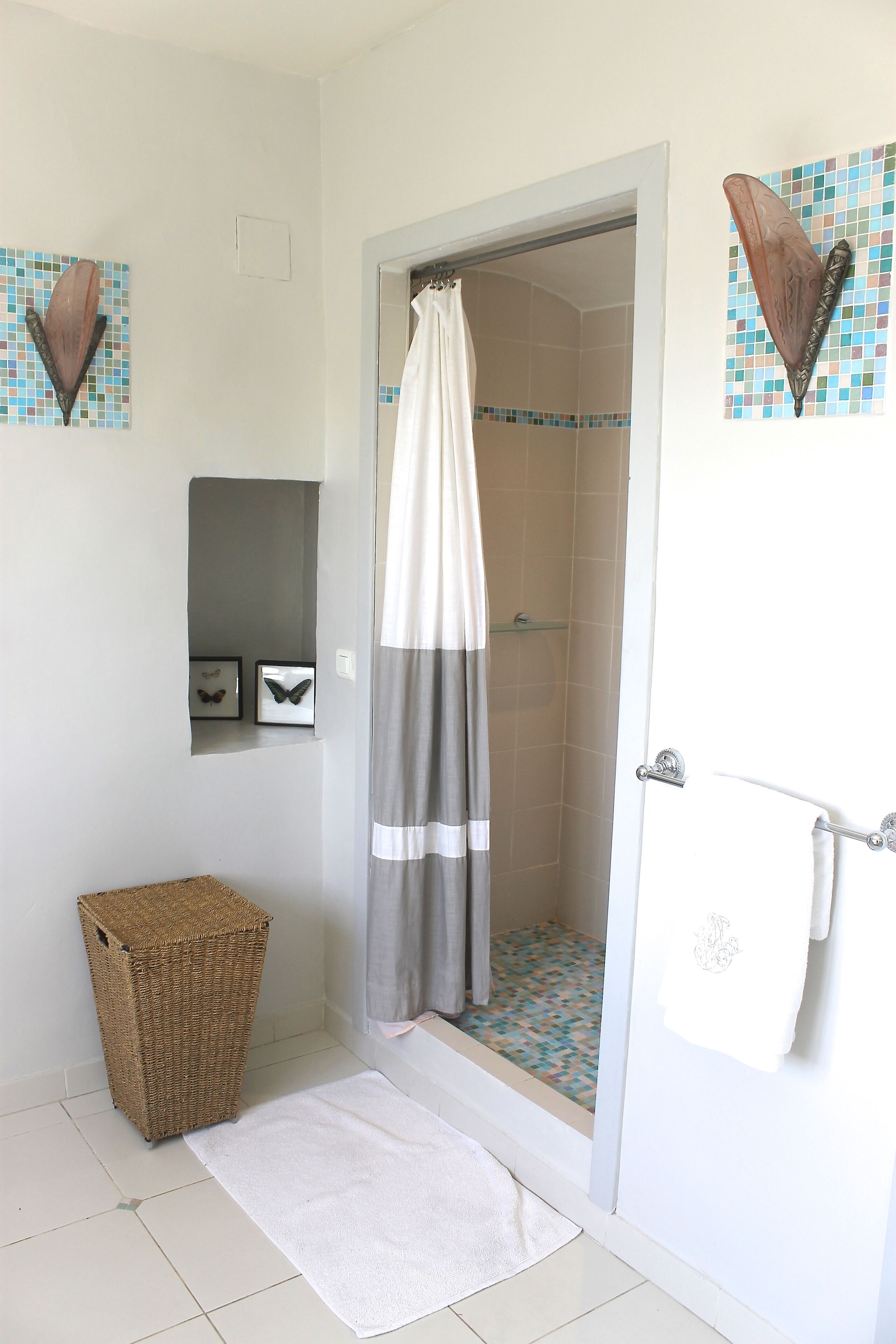 Un baño con ducha, un perchero para toallas, una cesta de mimbre y un pequeño estante con fotografías enmarcadas.