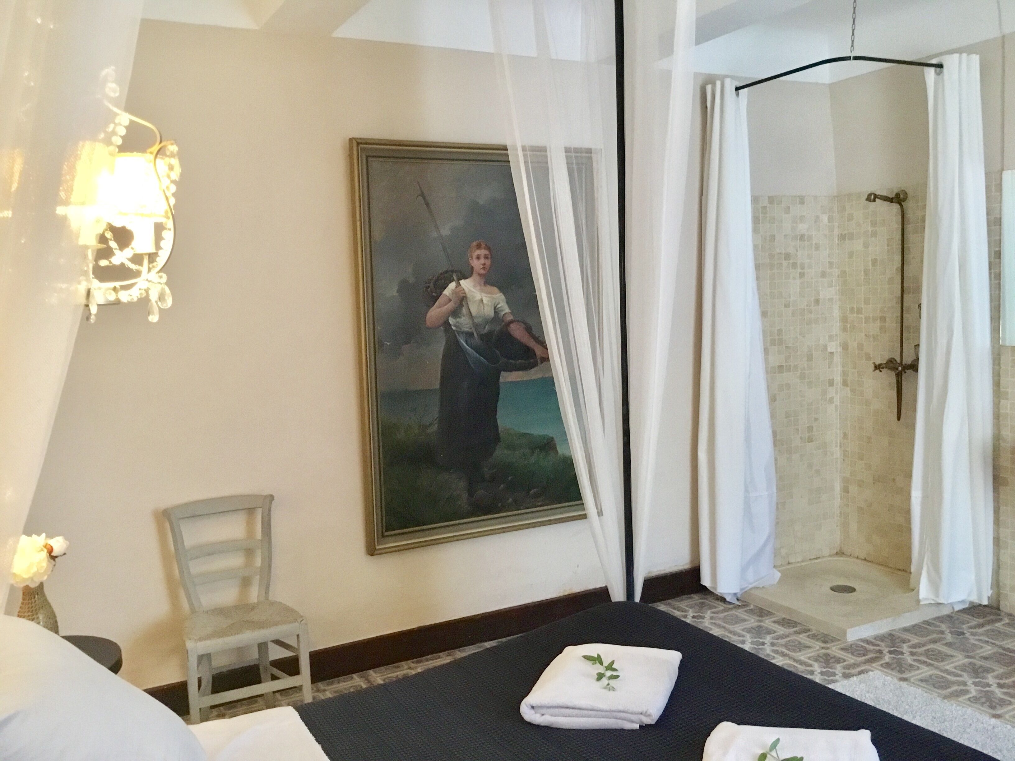 A hotel room with a bed, a chair, a painting, a shower, and a wall-mounted light fixture.