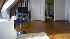 Een kamer met een schuine plafond, een houten vloer, een witte deur, een bed met een paarse deken, en een klein tafeltje met een lamp.