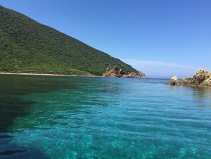 A coastal landscape with clear turquoise water, a green hill, and rocky outcrops.