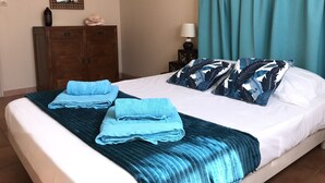 A neatly made bed with blue towels and tropical leaf-patterned pillows, a wooden wardrobe, a small table with a lamp, and teal curtains. - Caribbean villa on the beach (LE DIAMANT)