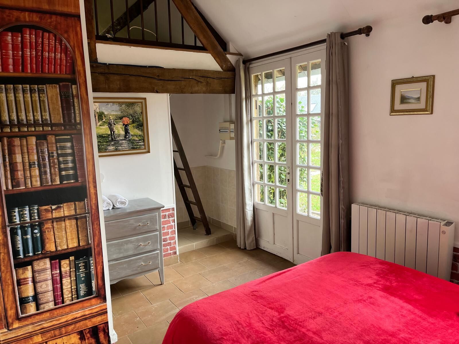 Une chambre avec une couvre-lit rouge, une bibliothèque en bois remplie de livres, un tableau accroché au mur et une fenêtre avec des rideaux.