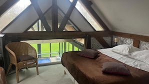 Un dormitorio con techo inclinado, una cama con almohadas, una silla de mimbre y vista a un césped verde.