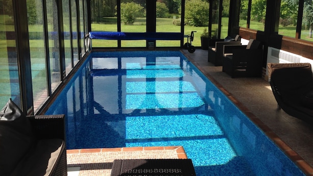 Una piscina interior con paredes de vidrio transparente, techo de madera y vistas a un paisaje verde.