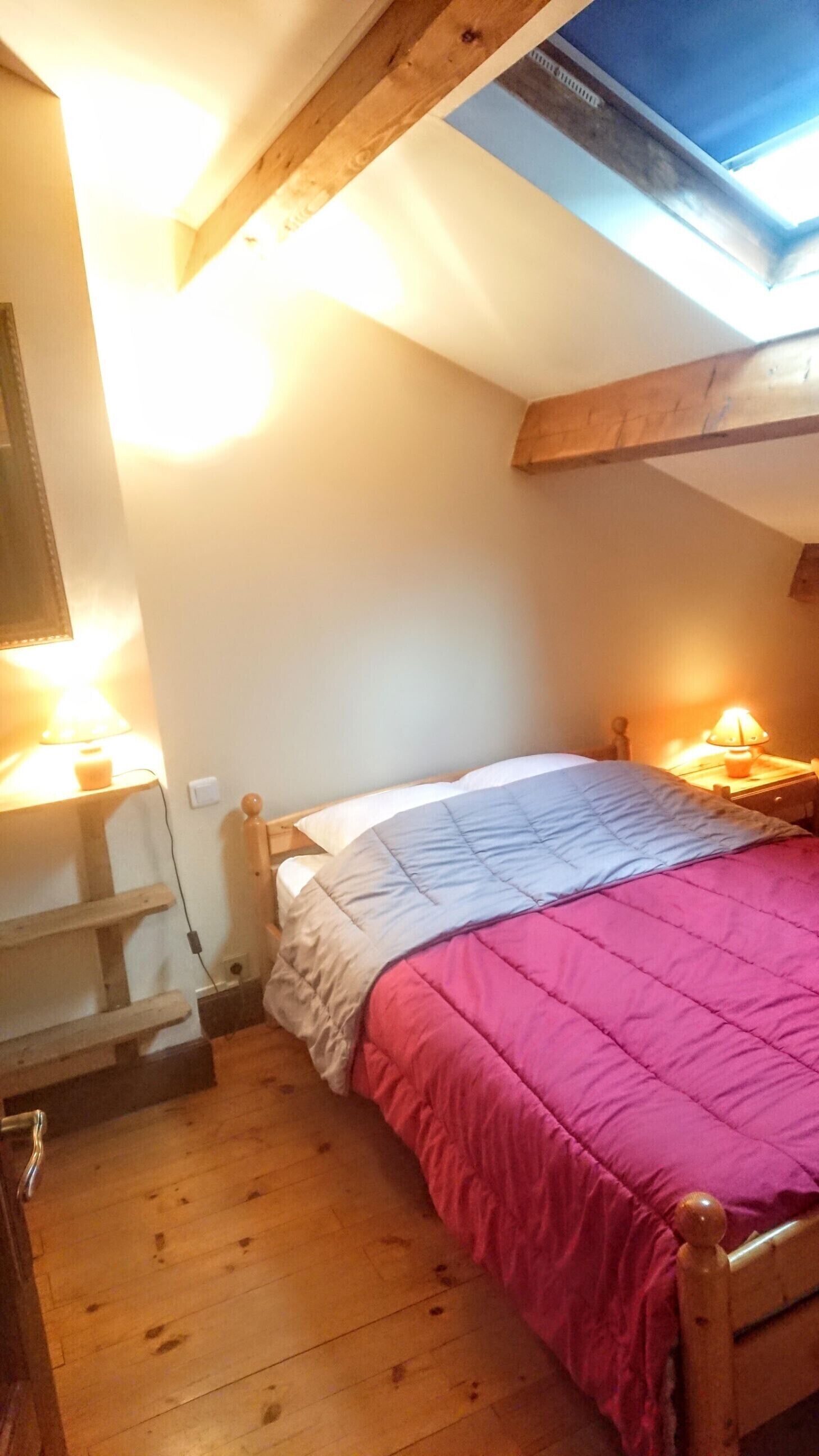 A bedroom with a sloped ceiling, a bed with a pink and blue quilt, two bedside lamps, a wooden bench, and a small table with a lamp.