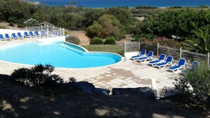 A swimming pool with blue lounge chairs, surrounded by greenery and a view of the sea. - New house in a touristy 3 stars residence in front of the sea (Belgodère)