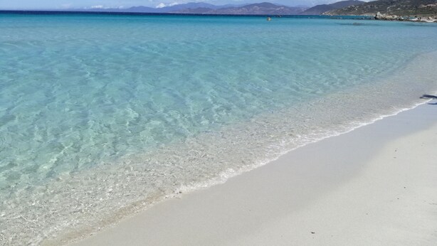 A beach with clear turquoise water, a sandy shore, and distant mountains under a clear sky. - New house in a touristy 3 stars residence in front of the sea (Belgodère)