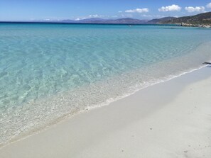 Een strand met helder turquoise water, een zandstrand en verre bergen onder een heldere lucht.