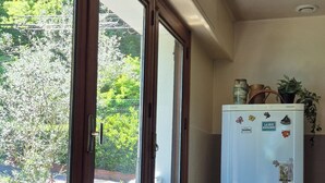 Eine Küche mit einem Kühlschrank, einem Fenster mit Blick auf Grünflächen, einem Tisch mit einer Obstkorb und einem Stuhl.