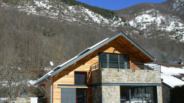 Una cabaña de montaña moderna con fachada de piedra, ventanas amplias y balcón de madera.