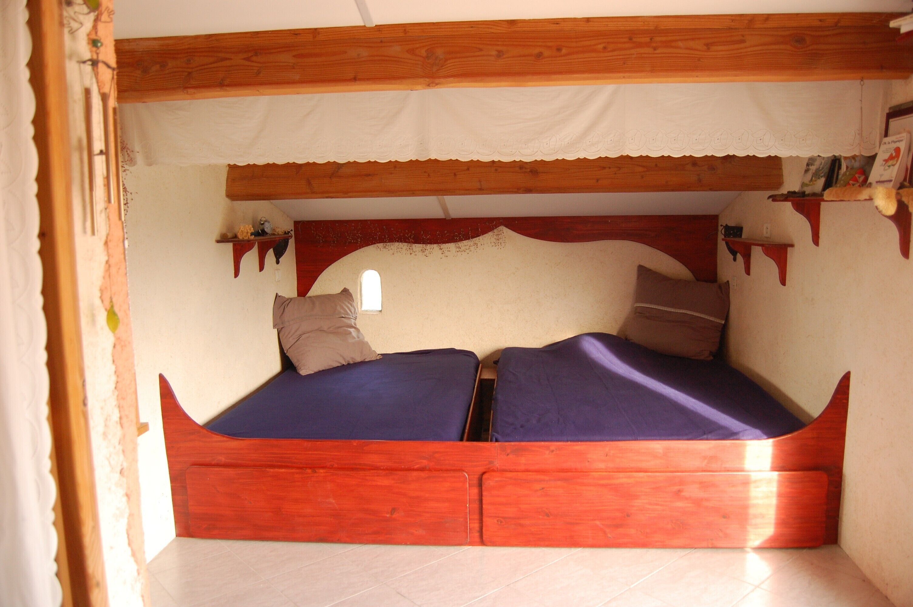 A small, cozy room with a wooden bed, a window, and a shelf with decorative items.