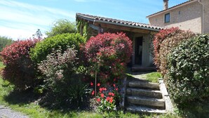 Un bâtiment rustique en pierre, doté d’un toit en tuiles, d’un escalier en pierre menant à l’entrée et d’une végétation luxuriante, notamment des arbustes à fleurs rouges.