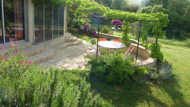 Une terrasse avec une table et des chaises, entourée d’une végétation luxuriante et agrémentée d’une pergola.