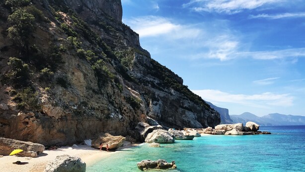 Ein Küstenstrand mit klarem türkisblauem Wasser, großen Felsen und einer felsigen Klippe.