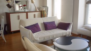 Una sala de estar moderna con un sofá blanco, dos almohadas moradas, una mesa de centro blanca y un sillón blanco.