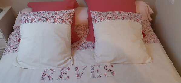 A bed with floral-patterned pillows and a white bedspread with the word "BEVERLY" embroidered in red and blue.