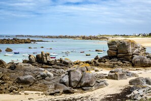 Una playa rocosa con barcos en el agua y casas al fondo.