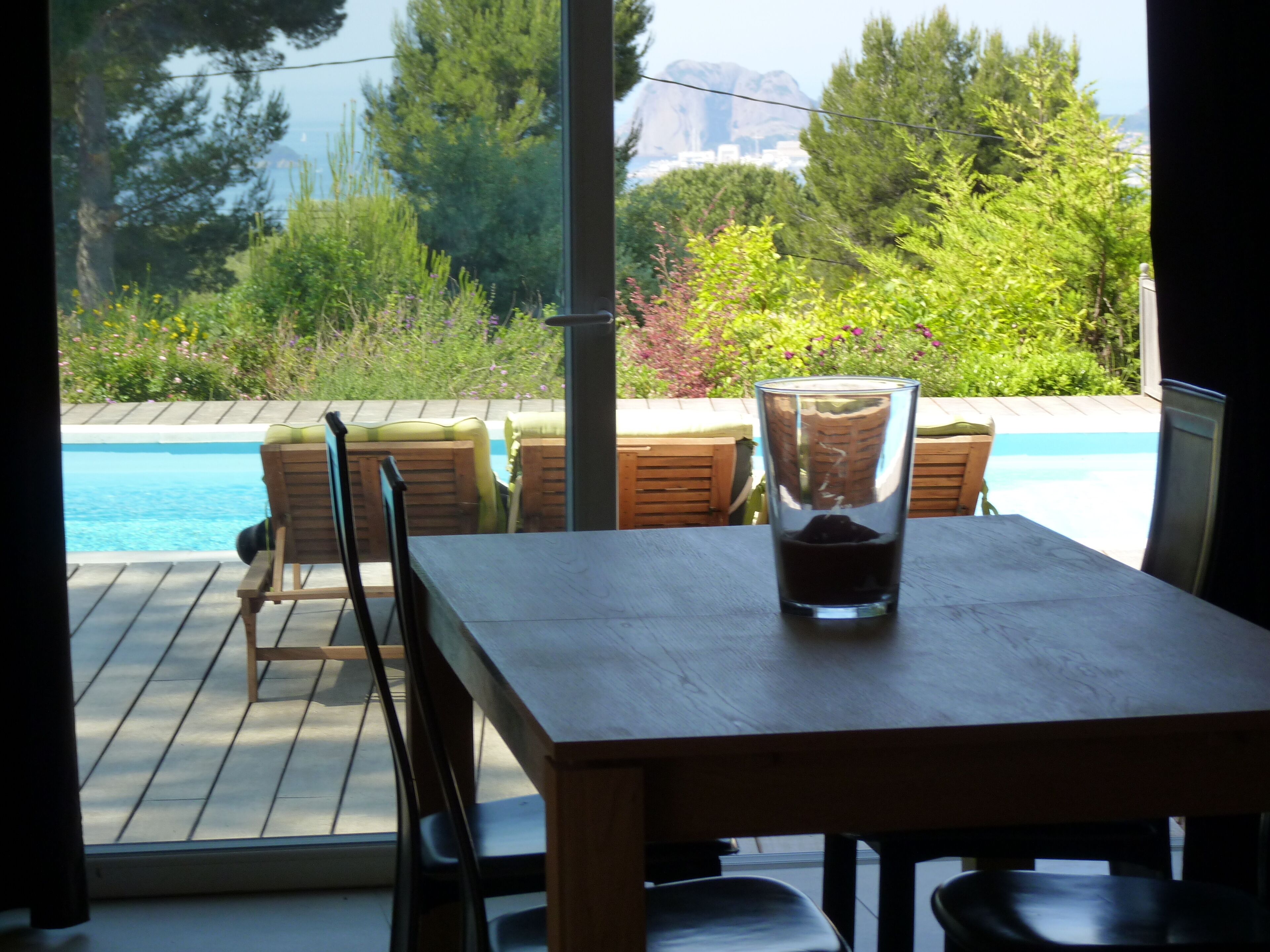 A wooden table with a glass and a chair in a room with a view of a swimming pool and greenery outside.