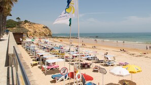 A beach with rows of sun loungers and umbrellas, a flagpole with a flag, and a rocky cliff in the background. - Ideal location, 20 m from the beach, in central Algarve, Albufeira (Albufeira)