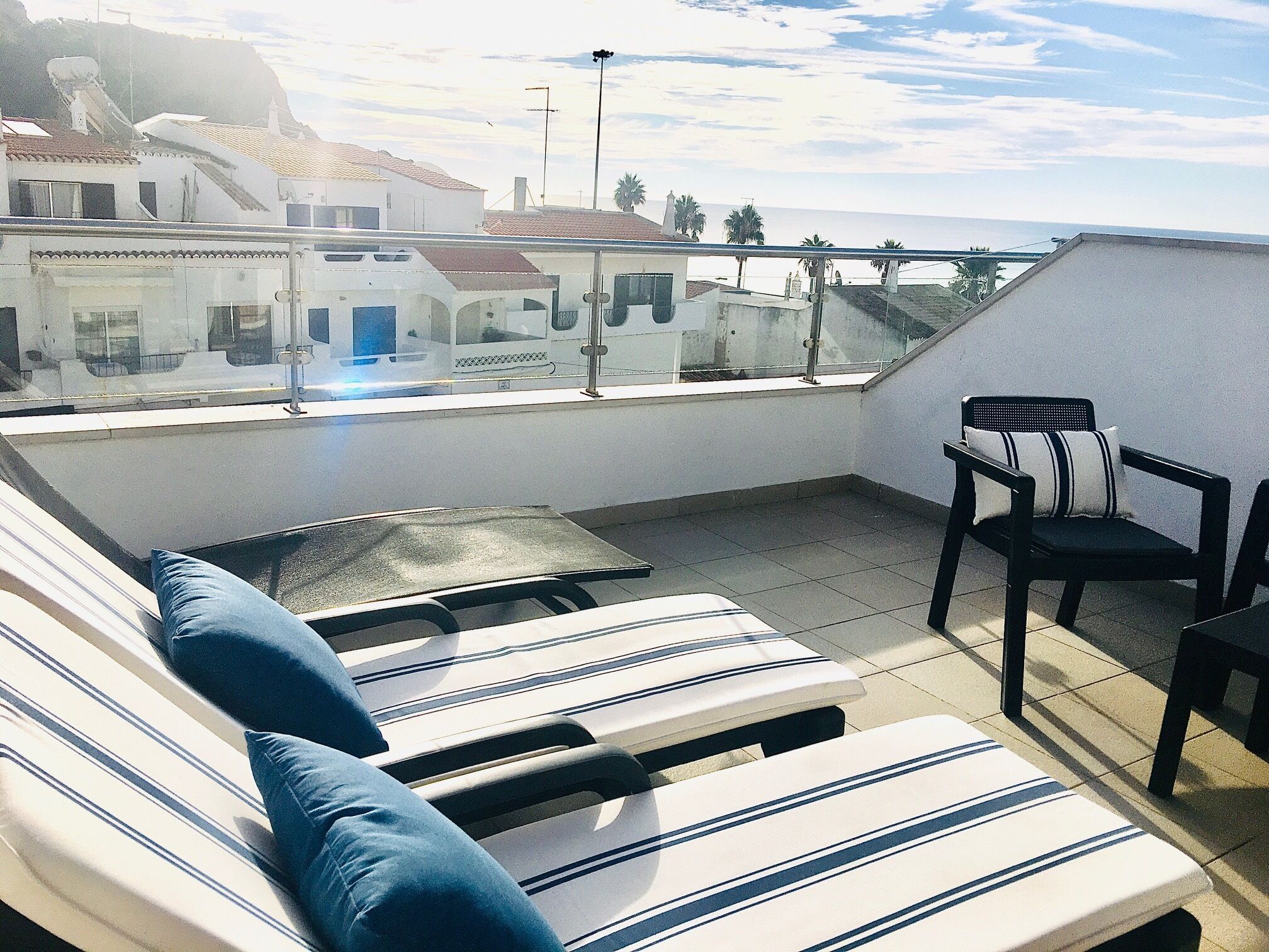 A rooftop terrace with lounge chairs and a table, overlooking a coastal town with buildings and a bridge.