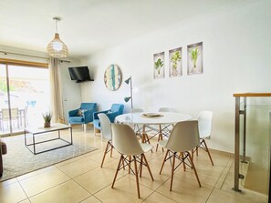 A modern living room with a dining table, blue armchairs, and a TV mounted on the wall.