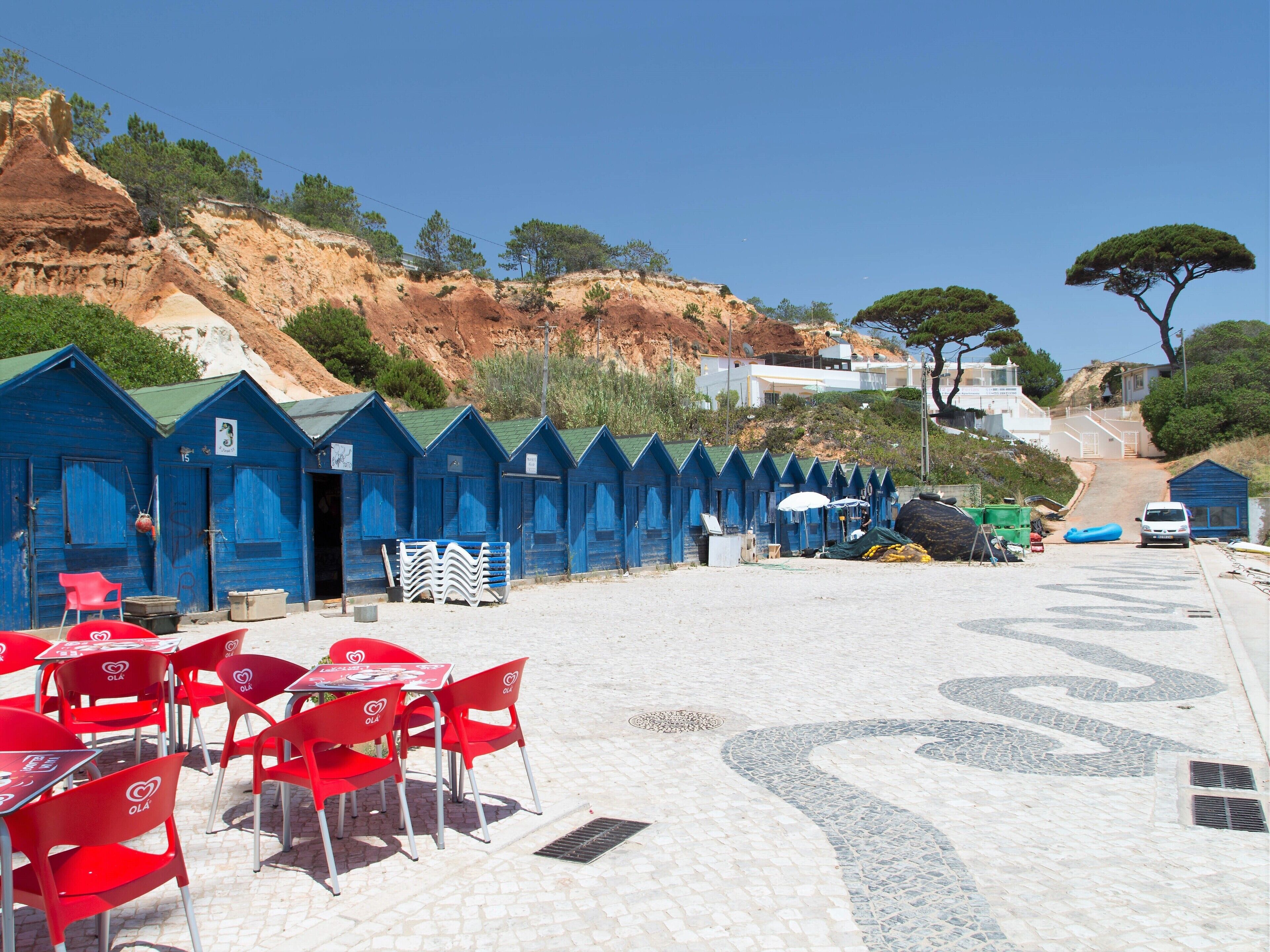 Een rijke rij blauwe strandhutten met een verharde looppad ernaartoe, rode stoelen en tafels op de voorgrond, en een helderblauwe lucht.