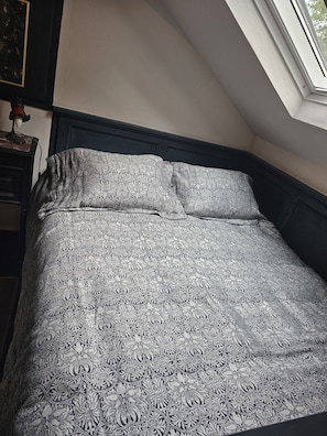 Un dormitorio con una colcha estampada, una mesita de noche con lámpara y una ventana con vistas a árboles.