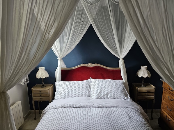 Une chambre à coucher avec un lit à baldaquin, une tête de lit rouge, une literie blanche à motifs, deux tables de chevet avec des lampes et une commode en bois.