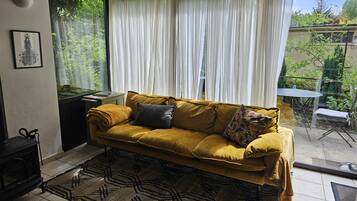 A living room with a yellow sofa, a patterned rug, and a view of a garden through sheer curtains.