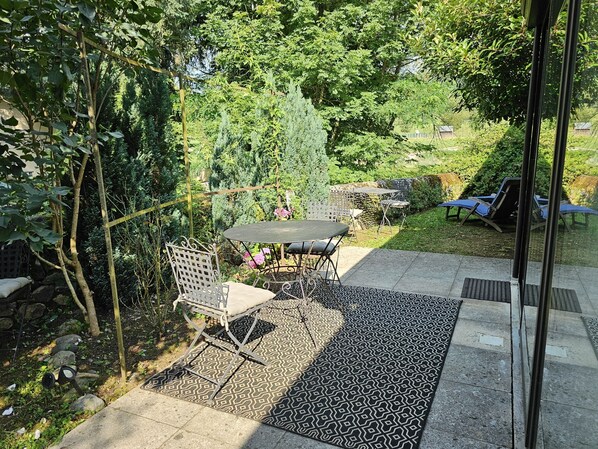A patio with a table and chairs, surrounded by lush greenery and a patterned rug. - Au Bord de la Rivière, Charming gîte with private (Saint Geniez D'olt Et D'aubrac)