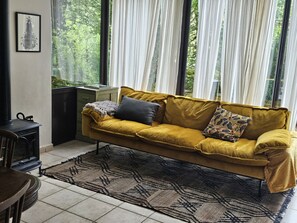 Une pièce avec un canapé jaune moutarde, un tapis à motifs et une vue sur la verdure à travers des rideaux transparents.