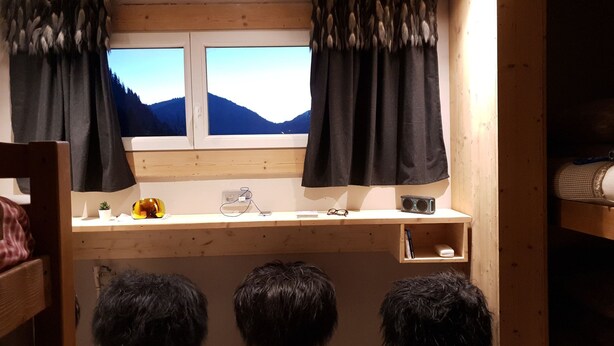 Une pièce avec un bureau en bois, deux petits tabourets noirs à poils longs, une fenêtre donnant sur les montagnes et une étagère avec une radio et des lunettes.