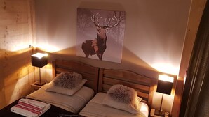 Une pièce chaleureuse aux murs en bois, avec deux lits, chacun agrémenté d’un oreiller moelleux et d’une lampe. Un tableau représentant un cerf est accroché au mur.
