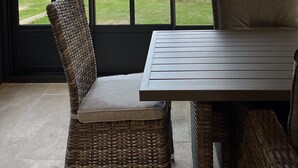 A room with a large window, wicker furniture, and a view of a grassy outdoor area with a wooden table and chairs.