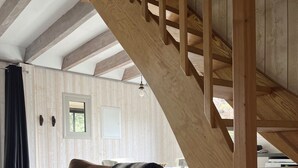 A wooden staircase leading to a loft bedroom with a large window, a brown leather sofa, and a black and white striped throw blanket.