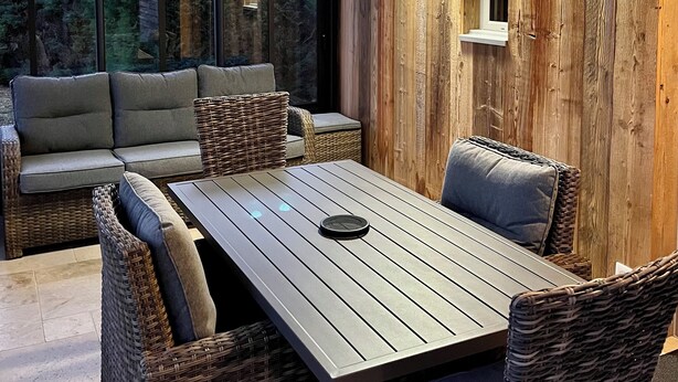 A modern outdoor seating area with a wooden table and wicker chairs, a cushioned sofa, and a view of the outdoors through large windows.