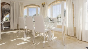 A dining area with a glass table and white chairs, large windows with curtains, and a view of the outdoors.