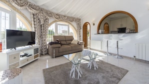A spacious living room with a glass coffee table, a sofa, a TV, and a kitchen area visible through an archway.