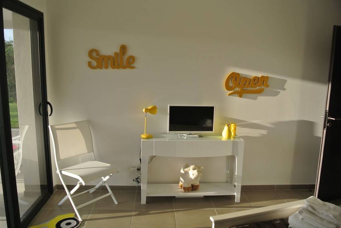 Une pièce avec un bureau blanc, une télévision et une étagère sur laquelle est posé un jouet en peluche. Il y a deux panneaux accrochés au mur, l’un porte l’inscription « Souriez » et l’autre « Ouvert ».
