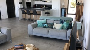 Una sala de estar moderna con un sofá gris, una mesa de centro de madera con libros y plantas, un televisor sobre un mueble de madera, y una cocina al fondo.