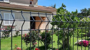 A house with a tiled roof, a garden with red flowers, and a metal fence in the foreground. - Small cozy nest for peaceful vacations (Lucciana)