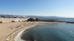A beach with people sunbathing and swimming, a stone wall, and boats in the background. - Bright, modern apartment in town center (Antibes)
