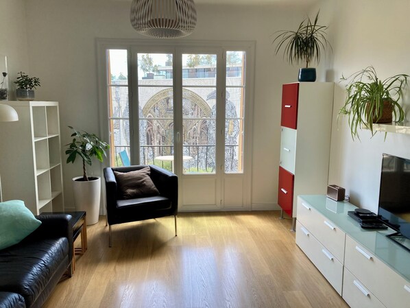 Un salon moderne avec un canapé en cuir noir, une table basse en verre, un fauteuil noir, une étagère blanche, un meuble rouge et blanc, un téléviseur et une grande fenêtre donnant sur un immeuble.
