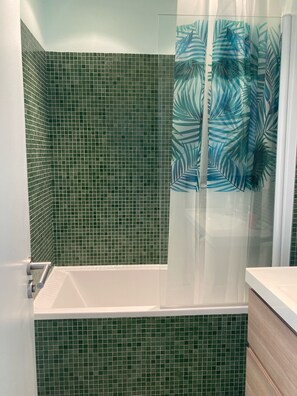 Une salle de bain avec des carreaux de mosaïque verts, une baignoire blanche et un rideau de douche orné d’un motif de feuilles.
