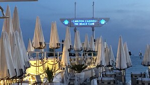 A beach club with rows of white umbrellas and lounge chairs, a sign indicating 'Carlton Cannes The Beach Club', and a view of the sea.