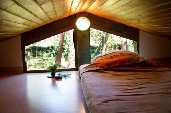Un dormitorio con techo inclinado y ventanas grandes, que ofrece vista a árboles y follaje exterior.