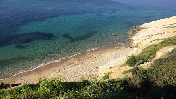 Paisatge costaner amb aigües cristal·lines, una platja sorrenca i penya-segats rocosos.