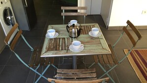 Zona de dinar amb taula preparada per a dues persones, incloent un bullidor, tasses i una cullera. Hi ha dues cadires de fusta i una banca. Es veuen una rentadora i un frigorífic al fons.